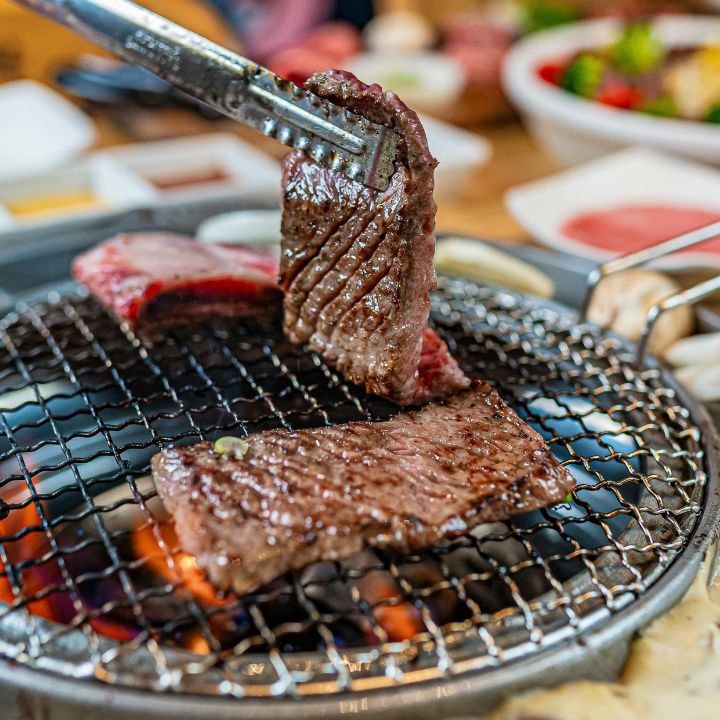 Korean-style barbecue, grilling thick beef cuts with side dishes in the background. 