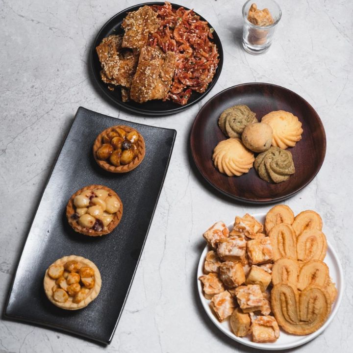 Various palmiers, nut tarts, cookies, Liuzhen almond cookies and seafood snacks on white dish against grey marble surface.