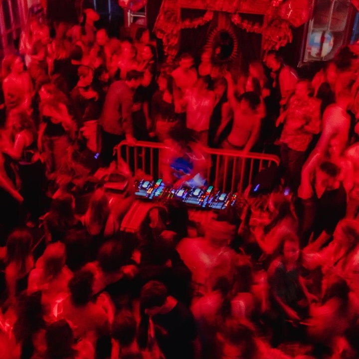 blurred image of dancers and clubbers at a nightclub surrounding a DJ
